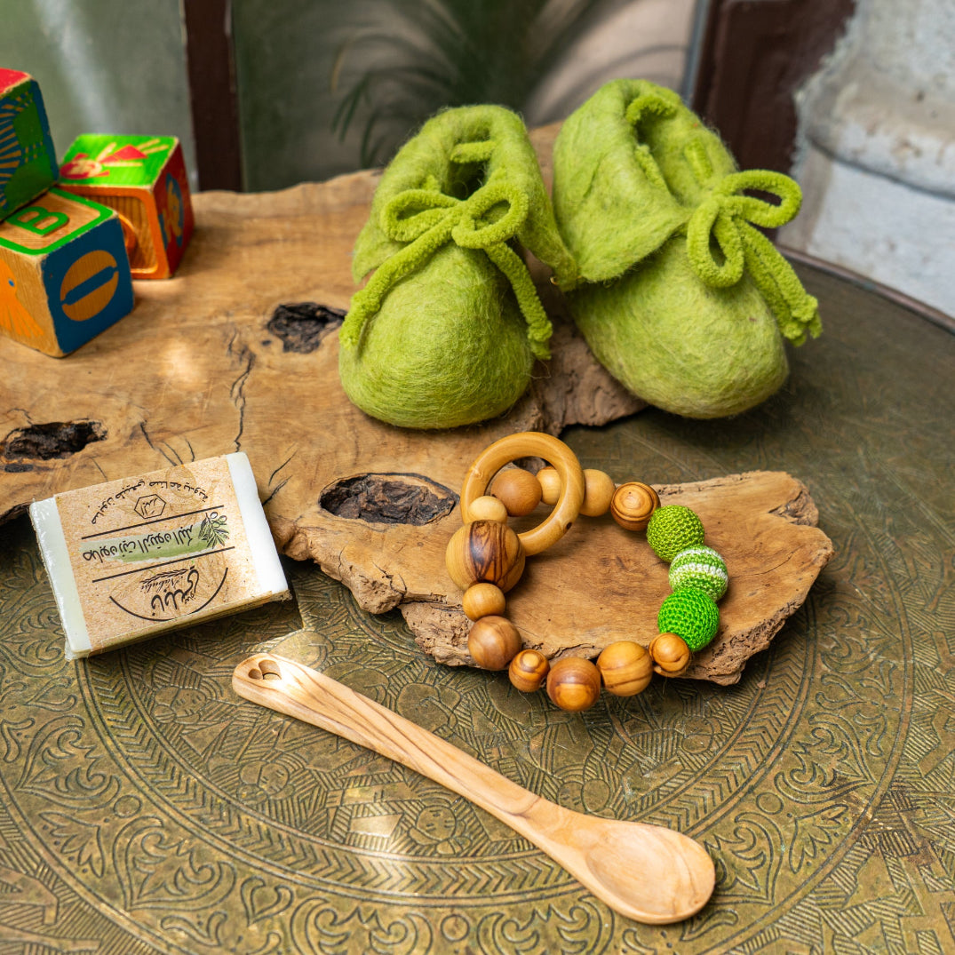 Green felted baby slippers on a wooden surface with a wooden spoon, soap, and toy in the background.