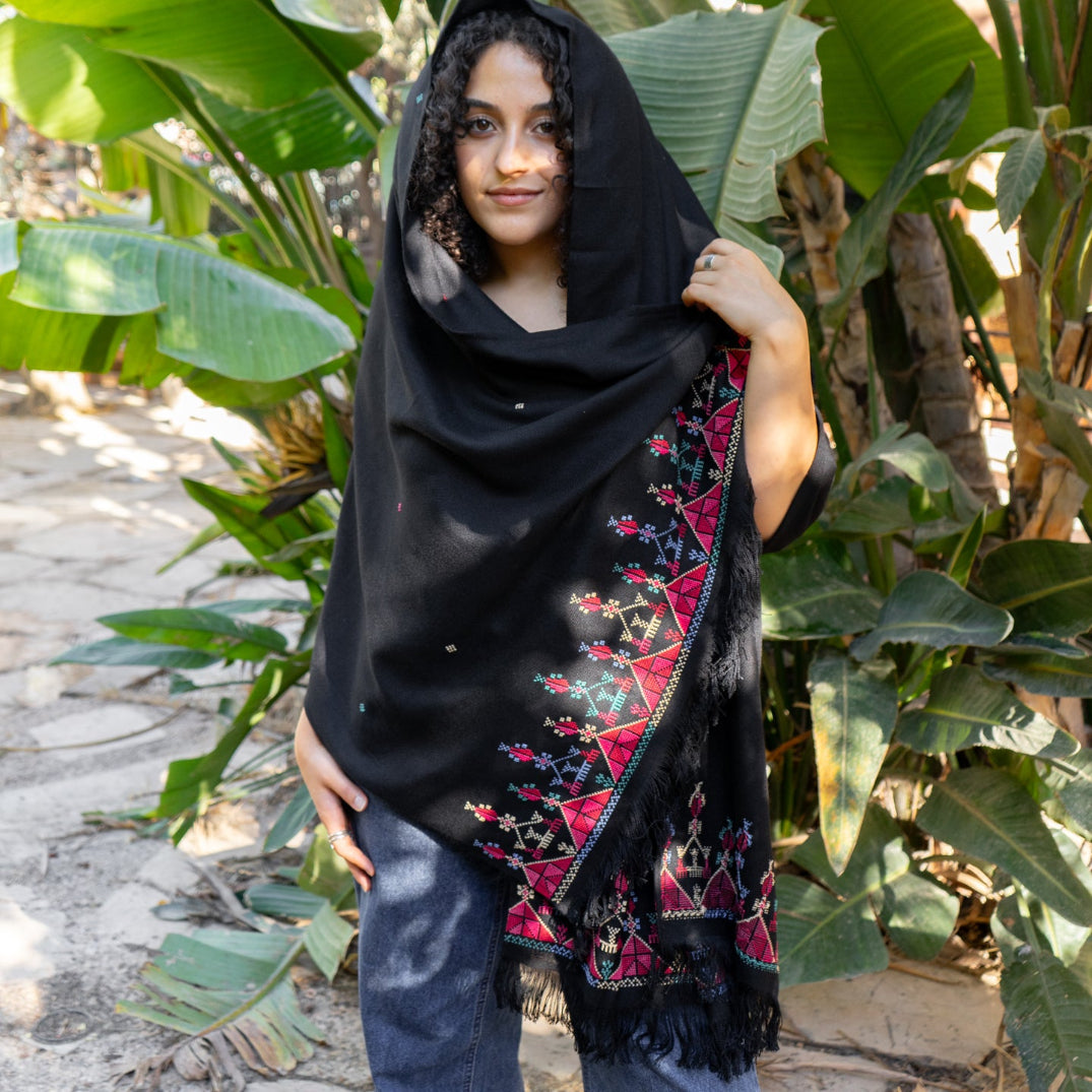 Woman wearing a black tatreez embroidered shawl in a natural setting with green plants.