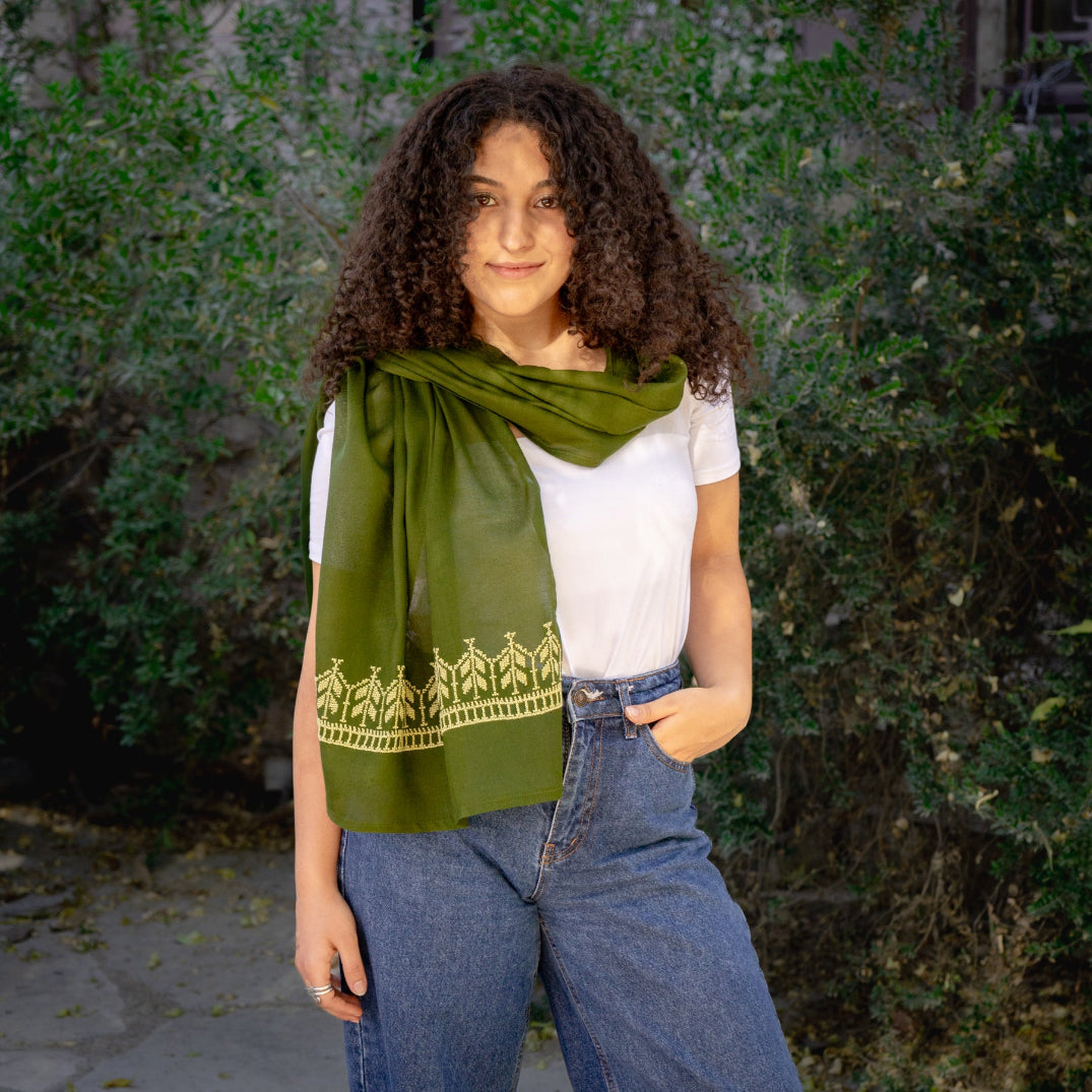 Palestinian woman wearing a green scarf with traditional tatreez, white shirt, and blue jeans standing outdoors.
