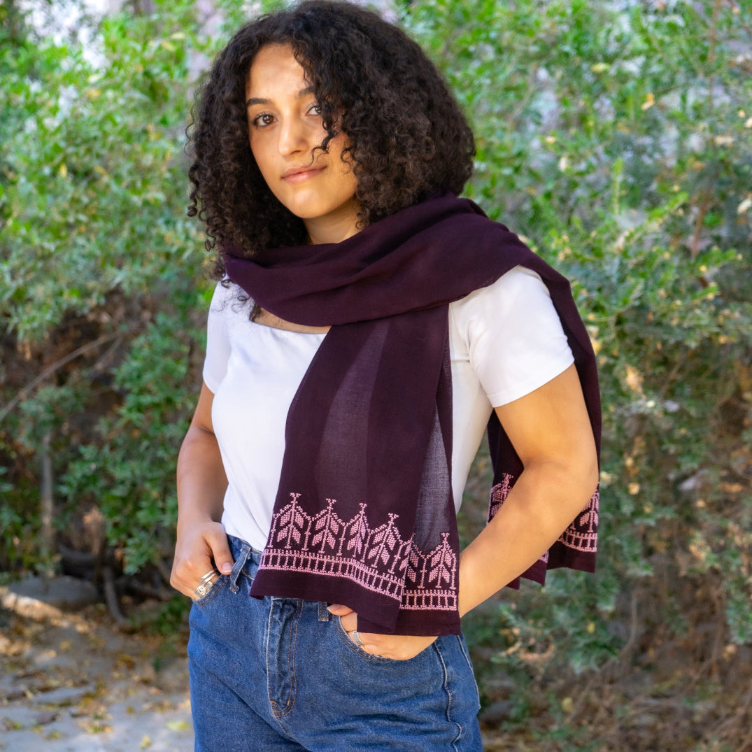 Woman wearing a dark purple summer scarf with a Palestinian tatreez, standing outdoors with greenery in the background