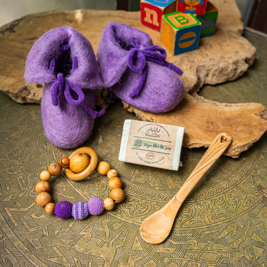 Purple handcrafted felted wool baby shoes on a wooden surface with a wooden spoon, beads, and soap bar.