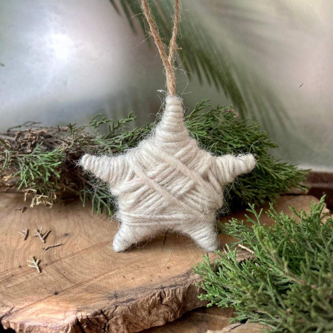 Hand spun wool Christmas ornament shaped like a star, with visible loops of wool and a twine hanging loop, displayed against a natural background with green foliage.