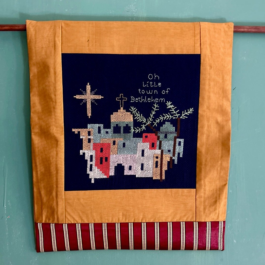 A handmade holiday wall hanging featuring the landscape of Bethlehem with a gold frame and traditional Palestinian embroidery, displaying text 'Oh little town of Bethlehem'.