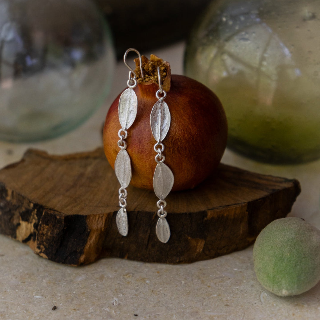Silver leaf-shaped earrings on a wooden block with a pomegranate and glass Hebron balls in the background.