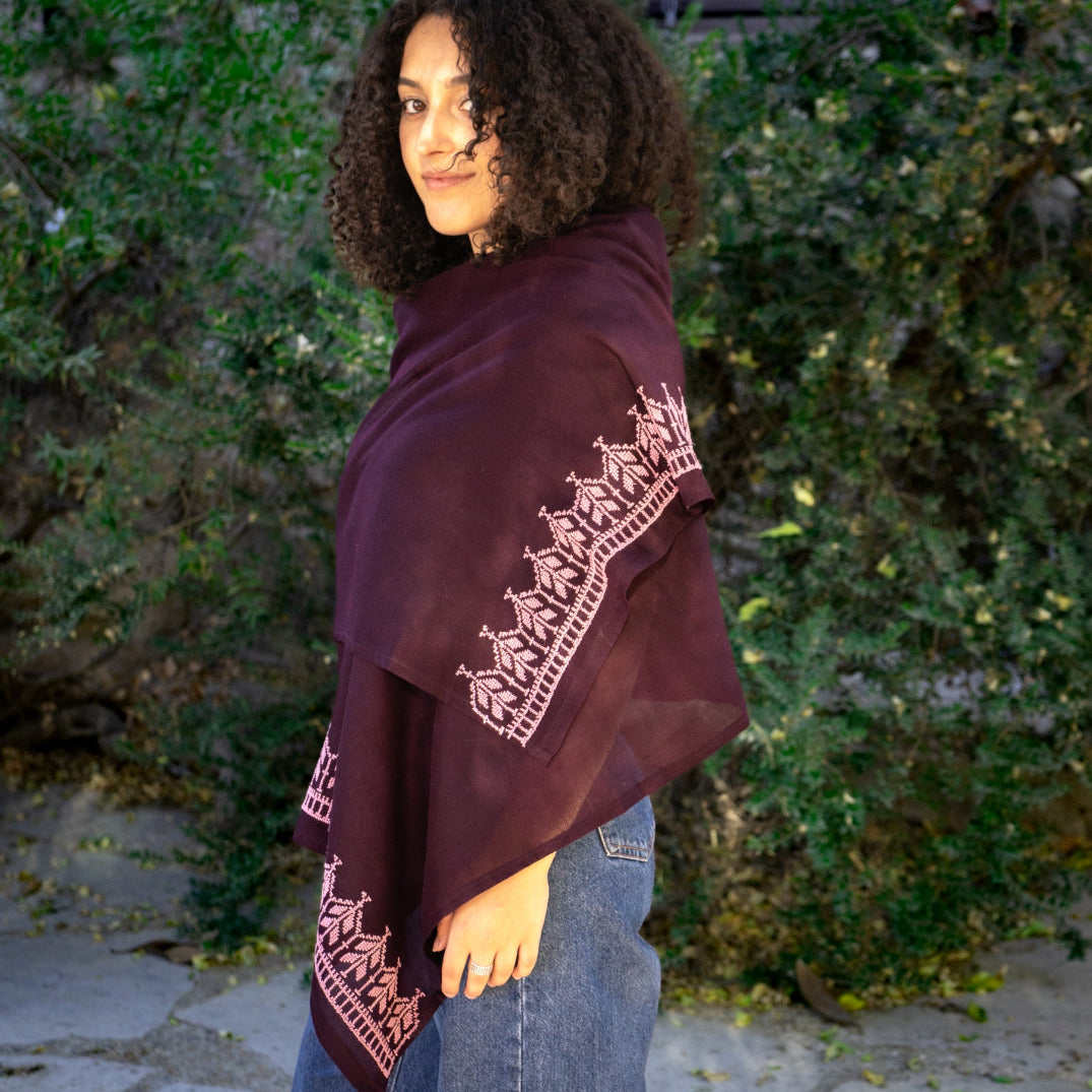 Palestinian woman wearing a dark purple shawl with lavender tatreez against a green bush background