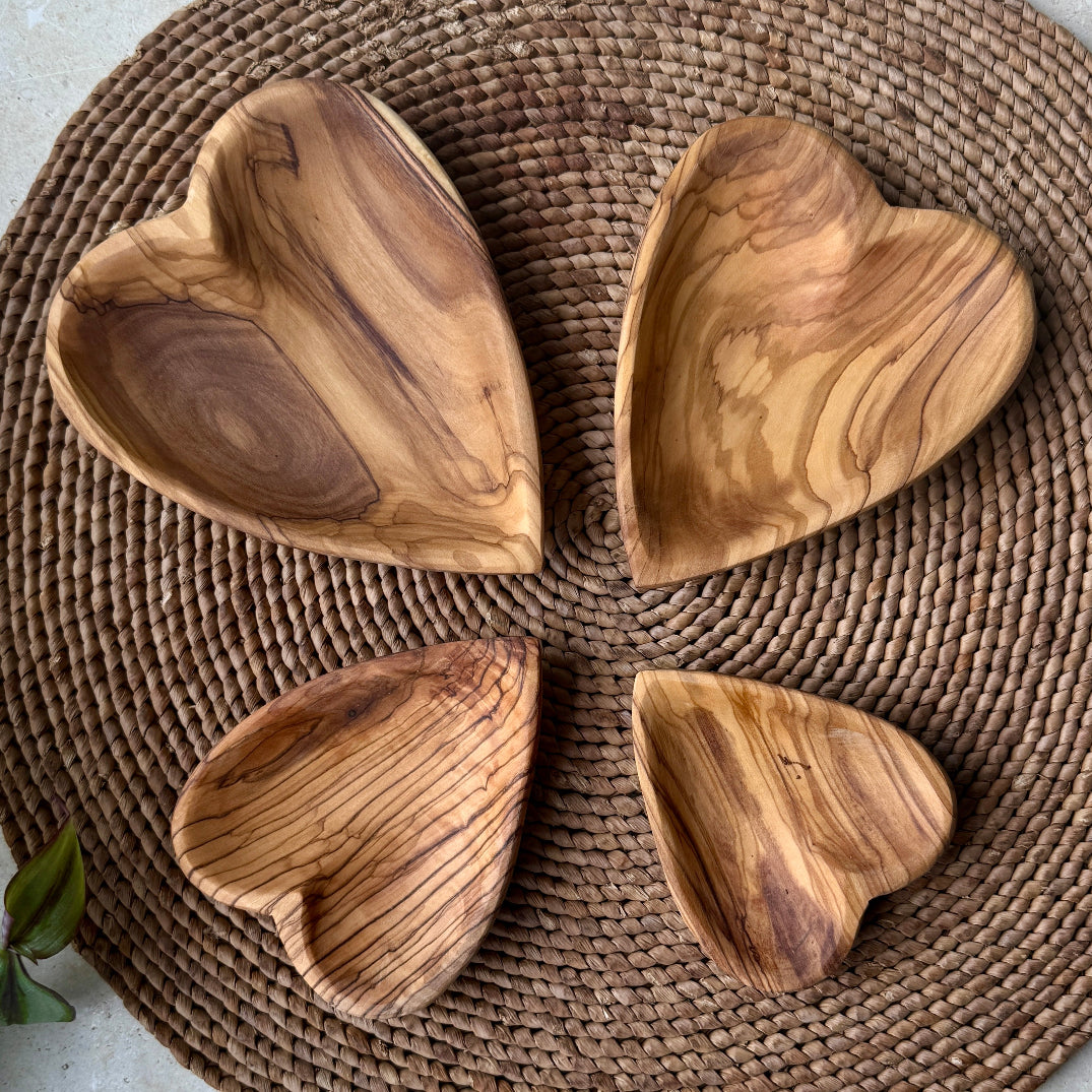 Four heart-shaped wooden serving dishes with natural olive wood grain patterns, arranged on a round woven brown placemat.